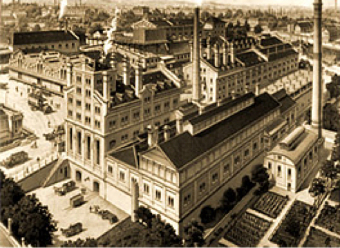 Historisches Fabrikgebäude mit vielen Fenstern, Schornsteinen und umgebenden Gärten.