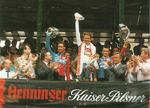 Drei Radfahrer feiern einen Sieg, der mittlere hebt einen Pokal, umgeben von Zuschauern.